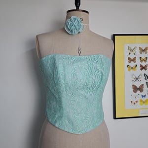 May include: A light blue lace corset top and matching choker with a flower accent. The corset is displayed on a mannequin. The top features a floral lace pattern. A framed butterfly print is visible in the background.
