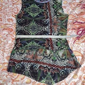 May include: A sleeveless top featuring a vibrant butterfly wing pattern in shades of green, brown, and purple. A white measuring tape is placed across the chest, showing a width of approximately 17 inches. The garment has a round neckline.