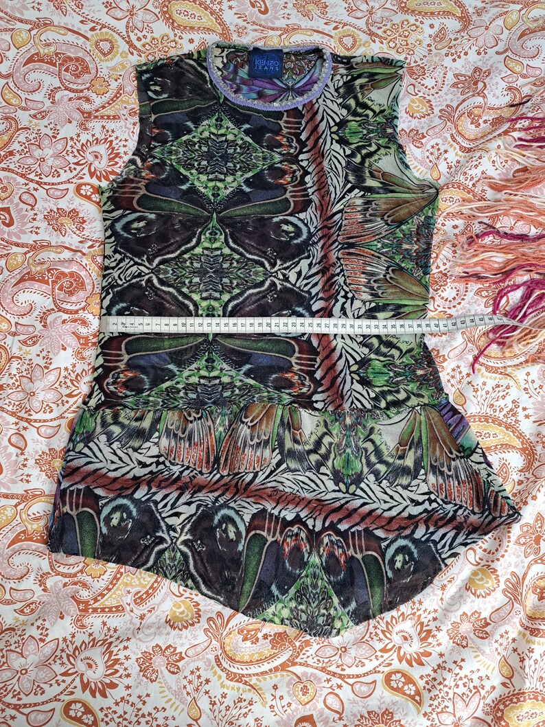 May include: A sleeveless top featuring a vibrant butterfly wing pattern in shades of green, brown, and purple. A white measuring tape is placed across the chest, showing a width of approximately 17 inches. The garment has a round neckline.