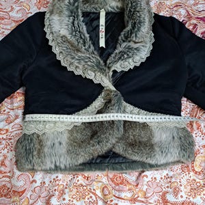 K&ouml;nnte beinhalten: Schwarze Bolerojacke mit Kunstfellbesatz und Spitzenkragen. Die Jacke hat einen einzelnen Knopfverschluss und wird mit einem Ma&szlig;band gemessen. Der Kunstfellbesatz der Jacke ist grau und braun.