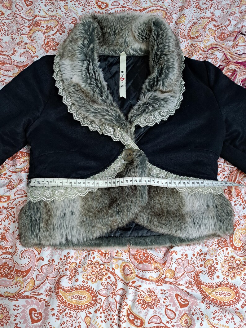 K&ouml;nnte beinhalten: Schwarze Bolerojacke mit Kunstfellbesatz und Spitzenkragen. Die Jacke hat einen einzelnen Knopfverschluss und wird mit einem Ma&szlig;band gemessen. Der Kunstfellbesatz der Jacke ist grau und braun.