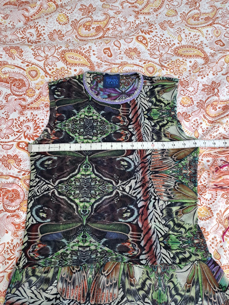 May include: A sleeveless Kenzo Jeans top featuring a vibrant, symmetrical butterfly wing print in shades of green, black, and brown. A white measuring tape across the chest indicates a width of approximately 16 inches. The neckline has a purple trim.