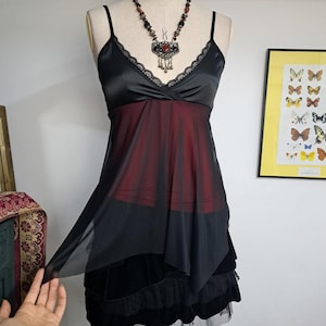 Puede incluir: Una camisola negra con una capa inferior de color rojo oscuro, con escote en V con ribete de encaje y tirantes finos. La prenda tiene una superposición asimétrica transparente y se combina con una falda negra de capas. Collar de cuentas.