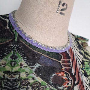 May include: A top featuring a vibrant, multi-colored graphic print with geometric and organic patterns in shades of green, black, and orange. The neckline is finished with a lavender-colored trim adorned with a row of small, metallic silver sequins.