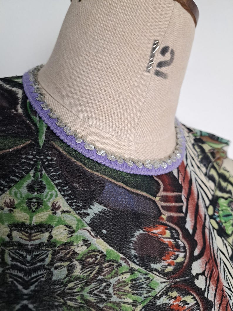 May include: A top featuring a vibrant, multi-colored graphic print with geometric and organic patterns in shades of green, black, and orange. The neckline is finished with a lavender-colored trim adorned with a row of small, metallic silver sequins.