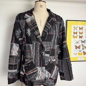 Könnte beinhalten: Ein schwarzer Blazer mit Patchwork-Design aus verschiedenen grauen und silbernen strukturierten Stoffbahnen, aufwendigen Nähten und Blumenstickereien. Die Jacke hat ein steigendes Revers, lange Ärmel und drei dunkle Knöpfe an der Vorderseite.