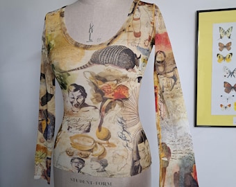 May include: A long-sleeved, sheer top with a scoop neck, featuring a collage of illustrations in muted tones. The design includes images of an armadillo, portraits, and botanical elements. The top is displayed on a mannequin.