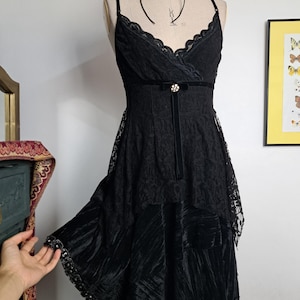 Puede incluir: Un vestido negro sin mangas con corpiño de encaje, escote en V, tirantes finos y una cinta de terciopelo en la cintura con un pequeño adorno de pedrería circular. La falda es de tela plisada y texturizada con una capa de encaje.