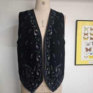 Può includere: Un gilet nero senza maniche con un profondo scollo a V e un intricato ricamo di paillettes blu scuro e iridescenti in un motivo paisley. Il capo è decorato con paillettes sparse sui pannelli frontali e un orlo a punta.