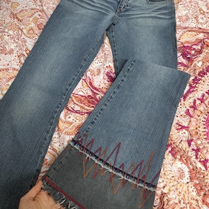 Puede incluir: Vaqueros azules con diseño de pernera acampanada. Los vaqueros presentan costuras decorativas rojas, naranjas y azules en la parte inferior de la pernera, con un dobladillo deshilachado. Los vaqueros tienen un diseño clásico de cinco bolsillos y cierre de botón y cremallera.