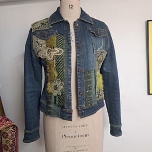 May include: A blue denim jacket with decorative patches of lace, faux fur, and embroidered details. The jacket has a classic collar, button closures, and two chest pockets. The patches include shades of green, cream, and blue.