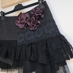 May include: A dark gray skirt with black lace and tulle detailing, adorned with three dark purple fabric flowers. The skirt is hanging on a black hanger against a light wood background.