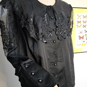 May include: Black blouse with a decorative collar and sheer sleeves. The collar is embellished with sequins and beads. The sleeves have a sheer overlay with lace detailing. The blouse has button closures and is displayed on a mannequin.