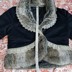 K&ouml;nnte beinhalten: Schwarze Bolerojacke mit Kunstfellbesatz und Kunstfellkragen. Die Jacke hat eine Spitzenborte an den Kanten und einen einzelnen Knopfverschluss. Ein Ma&szlig;band ist auf der Jacke platziert.