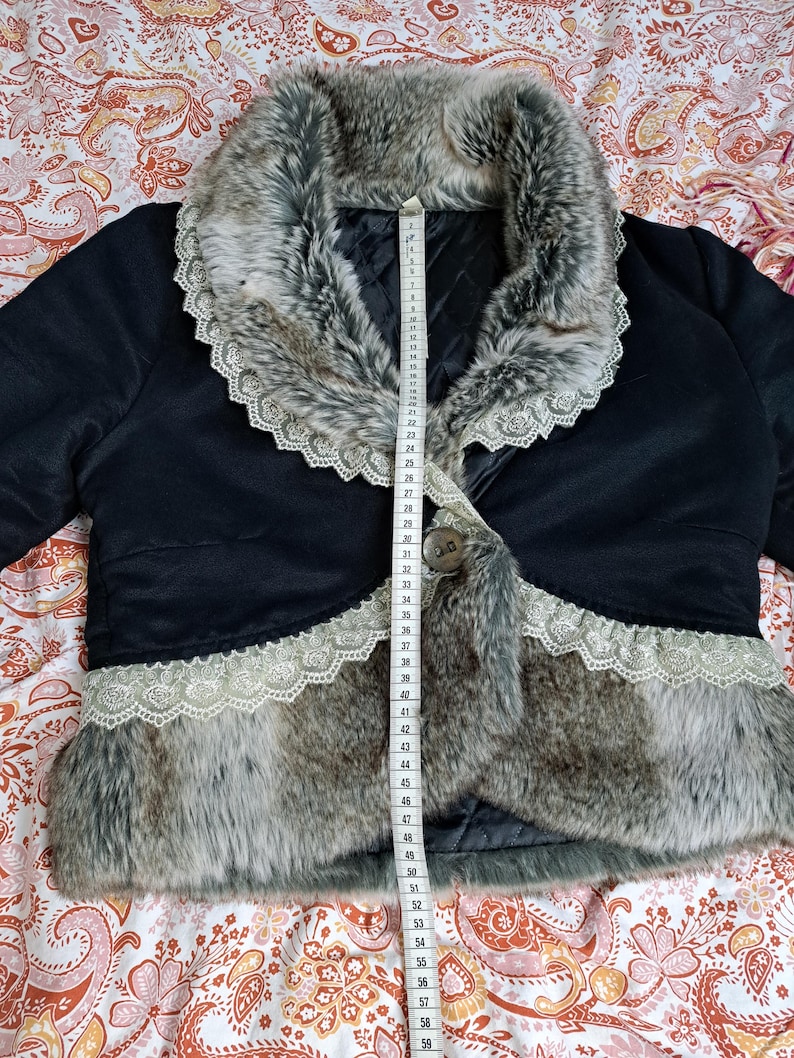 K&ouml;nnte beinhalten: Schwarze Bolerojacke mit Kunstfellbesatz und Kunstfellkragen. Die Jacke hat eine Spitzenborte an den Kanten und einen einzelnen Knopfverschluss. Ein Ma&szlig;band ist auf der Jacke platziert.