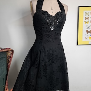 May include: A black halter dress with a floral pattern and a sweetheart neckline. The bodice is embellished with rosettes and sequins. The dress is displayed on a mannequin and has a flared skirt.