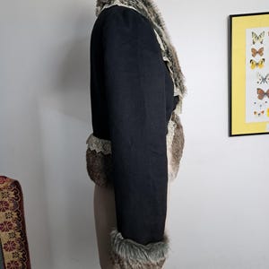K&ouml;nnte beinhalten: Schwarze Jacke mit Kunstfellbesatz an Kragen, Manschetten und Vorderseite. Die Jacke hat ein kurzes Design und eine Spitzenborte an den Kanten. Das Kunstfell ist eine Mischung aus Braun, Grau und Wei&szlig;.
