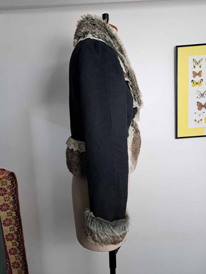 K&ouml;nnte beinhalten: Schwarze Jacke mit Kunstfellbesatz an Kragen, Manschetten und Vorderseite. Die Jacke hat ein kurzes Design und eine Spitzenborte an den Kanten. Das Kunstfell ist eine Mischung aus Braun, Grau und Wei&szlig;.