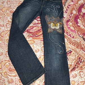 May include: Dark wash denim jeans with a decorative butterfly design on the front. The butterfly is embellished with sequins and beads. The waistband features a decorative embroidered trim. The jeans have a classic five-pocket design and a flared leg.
