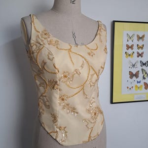 May include: A sleeveless cream-colored bodice featuring a scoop neckline and an intricate pattern of gold-toned embroidery, metallic thread, and clear sequins arranged in swirling floral and vine motifs across the front of the garment.