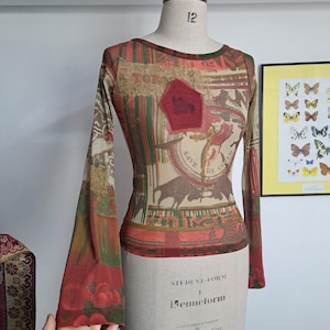May include: A long-sleeved, semi-sheer top featuring a multi-colored collage print with red, green, and brown tones. The design includes bull silhouettes, circular text reading 'SAVE THE QUEEN', and a red pentagonal patch on the chest area.