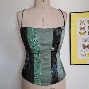 May include: A patchwork bustier top with thin straps, displayed on a mannequin. The design includes black, teal sequin, and sage green satin panels, with floral embroidery. The mannequin is labelled "Student-Form Kenneform".