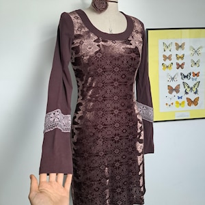 May include: A brown long-sleeve dress with a patterned bodice and lace accents on the sleeves. The dress is displayed on a mannequin with a matching choker. A framed butterfly print hangs in the background.