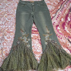 May include: Olive green denim jeans with flared bottoms. The jeans feature floral embroidery and fringed detailing on the front. The flared bottoms have a patterned mesh overlay. The jeans are laid flat on a patterned surface.