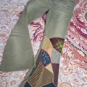 May include: Olive green corduroy flare pants featuring the word LOVE embroidered in black on the front waistband. The lower left leg is decorated with a large, multi-textured patchwork panel consisting of various fabric patterns and colors.