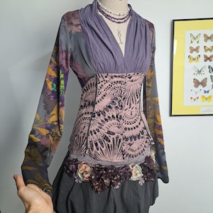May include: A unique dress with a purple bodice and long sleeves featuring a floral pattern. The dress has a pink, patterned midsection and a gray, gathered skirt. A pearl necklace and floral accents adorn the dress, displayed on a mannequin.