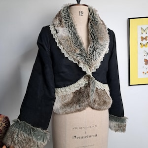 K&ouml;nnte beinhalten: Schwarze, kurze Jacke mit Kunstfellbesatz und -kragen. Die Jacke hat einen Knopfverschluss und Spitzendetails. Die Jacke wird auf einer Schneiderpuppe pr&auml;sentiert.