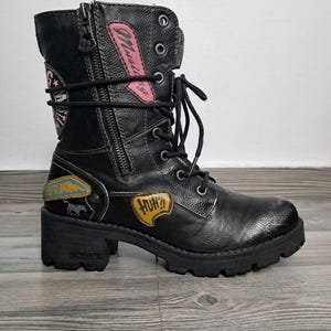 以下が含まれることがあります： 黒い革製のコンバットブーツ。サイドジッパー、レースアップクロージャー、チャンキーヒールが特徴です。ブーツには、ピンク色の「Mustang」という文字を含む、テキストとグラフィックのカラフルなパッチが付いています。ソールは厚く、ラグが付いています。