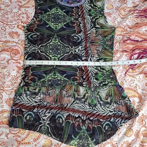May include: A sleeveless top with a multicolored butterfly wing pattern in shades of green, brown, and black. A measuring tape across the chest shows a width of approximately 16 inches. The garment features a round neckline and a flared hem.
