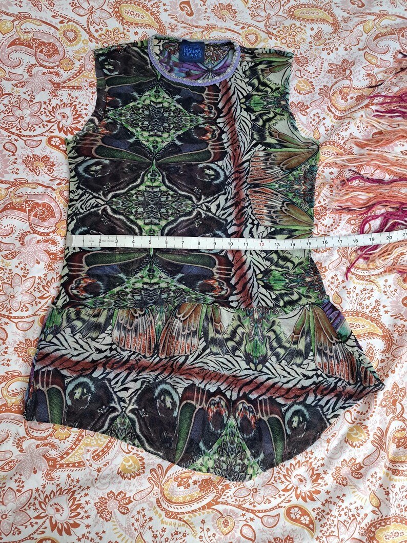 May include: A sleeveless top with a multicolored butterfly wing pattern in shades of green, brown, and black. A measuring tape across the chest shows a width of approximately 16 inches. The garment features a round neckline and a flared hem.