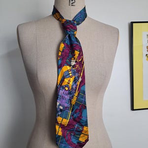 Puede incluir: Corbata de seda con un diseño detallado. La corbata presenta un patrón de cuadros verde azulado y amarillo con una ilustración central de una figura con atuendo amarillo y morado. La corbata está anudada y expuesta en un maniquí.