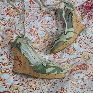 Op de afbeelding: Een paar sleehak sandalen met camouflageprint, kurken hakken en lange stoffen veters. De schoenen hebben gerafelde randen, een ronde neus en het woord Replay in rode letters op de zijkant van de kurken hak. De binnenkant is lichtgrijs.