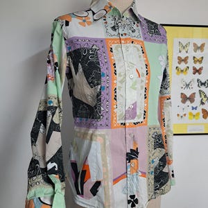 May include: A long-sleeve button-down shirt featuring a multi-colored patchwork design with various graphic patterns in shades of orange, purple, black, and light green. The shirt has a classic collar, a front button placket, and buttoned cuffs.
