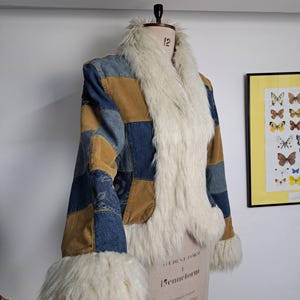 Puede incluir: Una chaqueta patchwork con adornos de mezclilla, pana y piel sintética. La chaqueta presenta un cuello y puños de piel sintética color crema, con paneles de mezclilla azul y pana marrón. La chaqueta se exhibe en un maniquí.