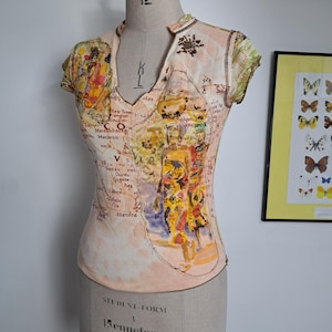 Könnte beinhalten: Ein kurzärmeliges Oberteil in blassem Pfirsichton mit einem aufgedruckten Kartenmotiv, das mit Illustrationen von Figuren in traditioneller Kleidung überlagert ist. Das Kleidungsstück hat einen V-Ausschnitt mit dunklen Perlen.