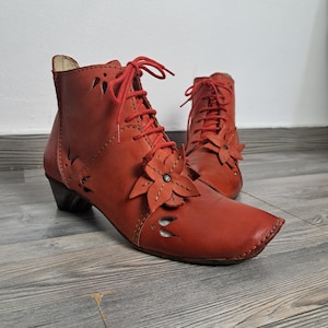 May include: A pair of reddish-orange leather ankle boots with floral cutouts and accents. The boots have a lace-up design with red laces, a small heel, and a flower applique on the side. The boots are displayed on a wooden floor.
