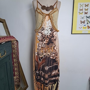 Op de afbeelding: Een mouwloze jurk tot op de grond met een mosterdgeel lijfje met ruches, een wit kanten paneel en een bruin rokgedeelte met een grafische print van rozen, franjes en een ketting met drie ronde bruine hangers.