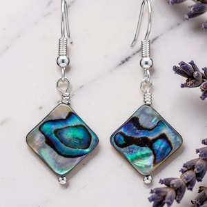 May include: Silver dangle earrings featuring diamond-shaped abalone shell pendants with iridescent blue and green hues. The earrings have a hook fastening and are shown with lavender sprigs.