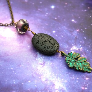 Könnte beinhalten: Eine Halskette mit einem Anhänger aus schwarzem Lavastein, einem grünen Blatt-Anhänger und einer violetten Glasperle. Die Kette ist aus Bronze.