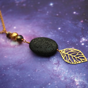 Könnte beinhalten: Eine goldene Kettenkette mit einem schwarzen Lavastein-Anhänger und einem goldenen Blatt-Anhänger. Die Kette liegt auf einem violetten Hintergrund mit goldenem Glitzer.