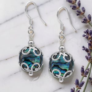 May include: A pair of silver earrings with iridescent abalone shell beads. The beads are encased in a decorative silver filigree design. The earrings hang from silver hooks. Lavender sprigs are in the background.