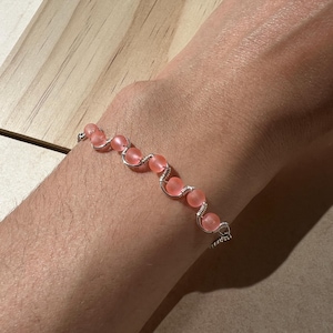 May include: A handmade bracelet featuring a series of pink, frosted glass beads strung together with silver-colored wire. The bracelet is designed to be worn on the wrist and has a delicate, feminine aesthetic. The beads are approximately 0.5 inches in diameter.