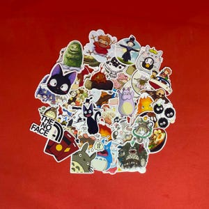 以下が含まれることがあります： 様々なキャラクターやデザインが描かれた、カラフルなアニメ風ステッカーのコレクション。 赤い背景に配置されたステッカーは、さまざまな形とサイズがあり、一部にはテキストが含まれています。