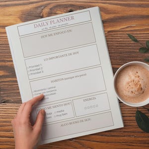 Op de afbeelding: Een dagplanner met de titel "DAILY PLANNER" in lichtpaarse letters. De planner heeft secties voor dagelijkse focus, prioriteiten, gewoonten en stemming. Een kopje koffie staat naast de planner op een houten oppervlak.