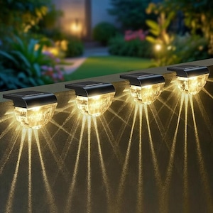 May include: Four solar-powered outdoor lights with a black top and a clear, textured glass globe. The lights are emitting a warm, yellow glow, casting light patterns on the surface below. The lights are mounted on a wall, with a blurred green background.