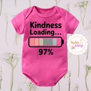 Peut inclure: Un body de bébé rose vif à manches courtes. Le body affiche le texte "Kindness Loading..." au-dessus d'un graphique de batterie avec des segments colorés et "97%" en dessous. Un logo de magasin pour bébés est visible à droite.